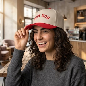 Könnte beinhalten: Beige Baseball Cap mit rotem Schirm und dem Wort "BOX" in roten Buchstaben. Die Kappe wird von einer lächelnden Person getragen. Der Hintergrund ist ein Café mit natürlichem Licht.