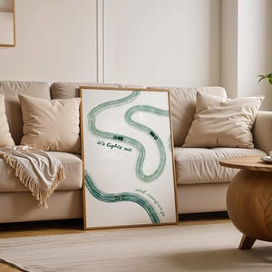 Può includere: Una stampa artistica incorniciata con un design di strada tortuosa verde su sfondo bianco. Il testo "It's lights out" e "and away we go" è stampato sull'opera d'arte. La stampa è esposta in un soggiorno con un divano beige.