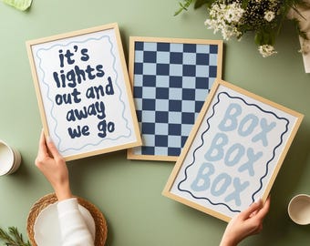 May include: Three framed art prints with light wood frames. One print has the text "it's lights out and away we go" in navy blue. Another has a blue and white checkerboard pattern. The third print says "BOX BOX BOX" in light blue.