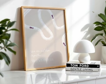 May include: A framed print with a beige background features a race track design with purple race cars. The quote "We win and lose together" by Sir Lewis Hamilton is printed on the artwork. A white mushroom-shaped lamp and books are in the foreground.