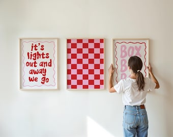 Op de afbeelding: Drie ingelijste kunstdrukken aan een witte muur. De prints hebben een rood-roze dambordpatroon en twee prints met de tekst "it's lights out and away we go" en "Box Box".