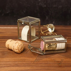 Puede incluir: Dos pequeñas cajas de vidrio con marcos plateados. Una caja está abierta y contiene un corcho de una botella de vino. La otra caja está cerrada y parece vacía.