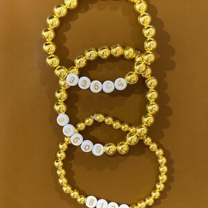 May include: Three gold beaded bracelets with white beads that have gold numbers on them. The numbers on the bracelets are 33604, 3609, and 813 with a small red heart.