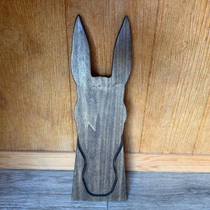 May include: A wooden donkey head-shaped wall decor piece. The donkey head is stained a dark gray color, with a black outline of the face and ears. The ears are long and pointed, and the face has a simple, stylized design. The decor is mounted on a wooden surface.