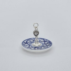 Könnte beinhalten: Eine kleine Schmuckschale aus Keramik mit weißem Untergrund und einem komplexen blauen Muster. Ein zentraler, silberfarbener Metallstab ragt aus der Mitte hervor, gekrönt von einem herzförmigen Anhänger und einem kleinen Ring.