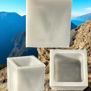 May include: Three white concrete square planters of different sizes. The planters are sitting on a rocky surface with a mountain range in the background.