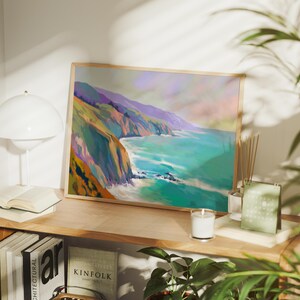 May include: A framed landscape art print depicts a coastal scene with cliffs, ocean, and sky in shades of teal, purple, and orange. The artwork is displayed on a wooden shelf with books, a lamp, and a candle.