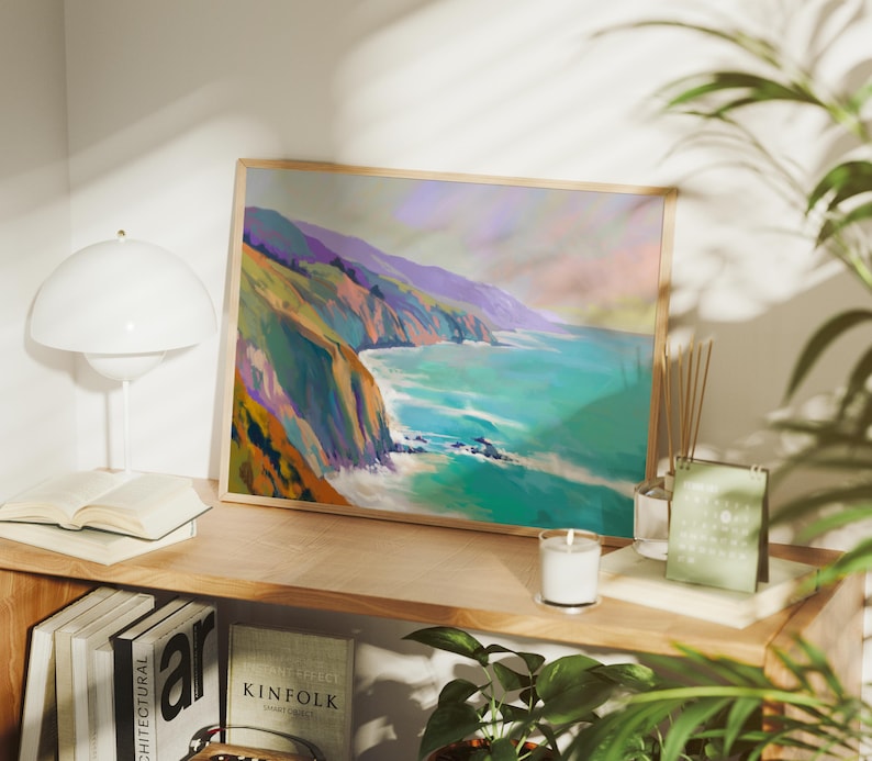 May include: A framed landscape art print depicts a coastal scene with cliffs, ocean, and sky in shades of teal, purple, and orange. The artwork is displayed on a wooden shelf with books, a lamp, and a candle.