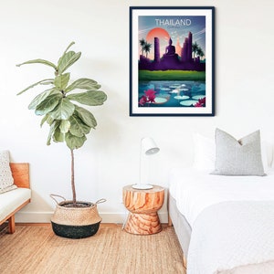 May include: A framed art print depicting a Thailand landscape with a sunset, a Buddha statue, and a lake with lily pads. The word "THAILAND" is printed at the top. The print is in a dark blue frame and hangs on a white wall.