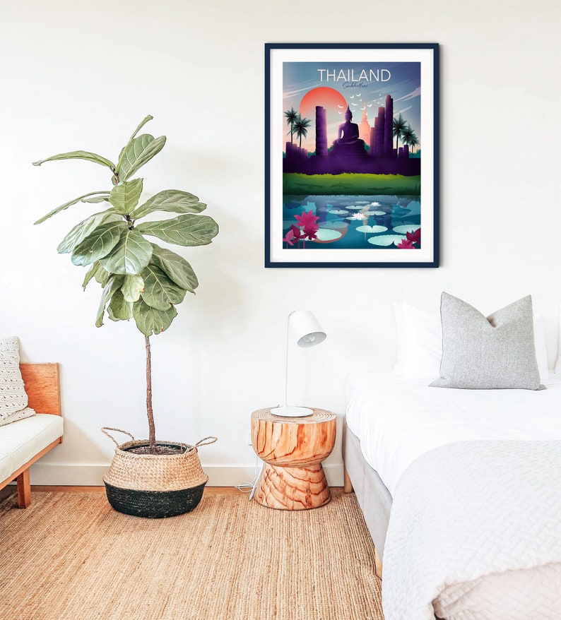 May include: A framed art print depicting a Thailand landscape with a sunset, a Buddha statue, and a lake with lily pads. The word "THAILAND" is printed at the top. The print is in a dark blue frame and hangs on a white wall.