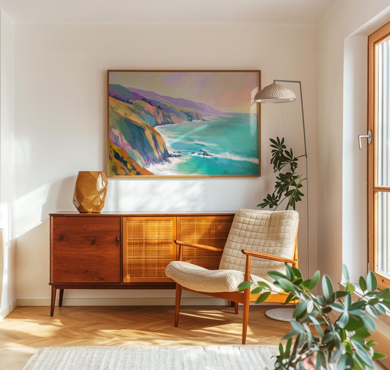 May include: A framed landscape painting depicts a coastal scene with turquoise water and colorful cliffs. A mid-century modern wooden cabinet with a geometric design and a beige armchair are in the room. A gold-colored vase sits on the cabinet.