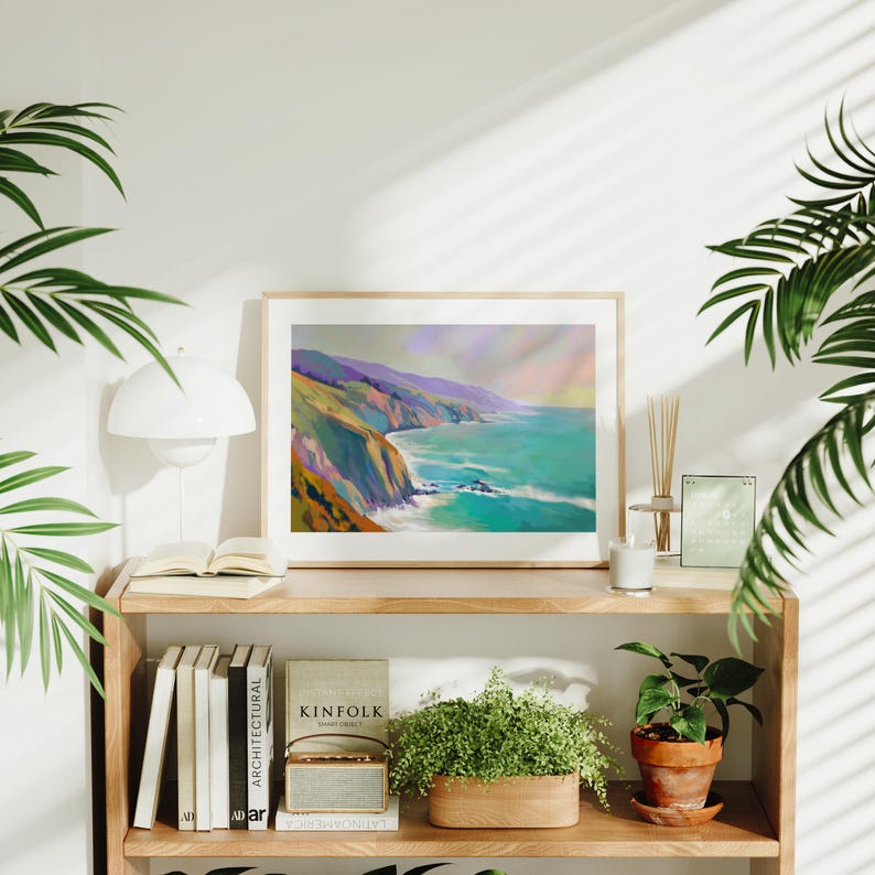May include: A framed landscape painting depicts a coastal scene with cliffs, turquoise water, and a colorful sky. The artwork is displayed on a wooden shelf with books, plants, and a lamp. The overall aesthetic is modern and serene, suitable for home decor.