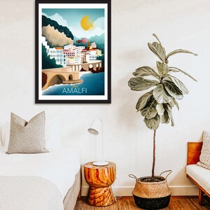 May include: Framed art print featuring a colorful illustration of Amalfi, Italy. The artwork depicts a coastal town with white buildings, a bridge, and the text "AMALFI" in white. The sky is blue with a yellow sun.