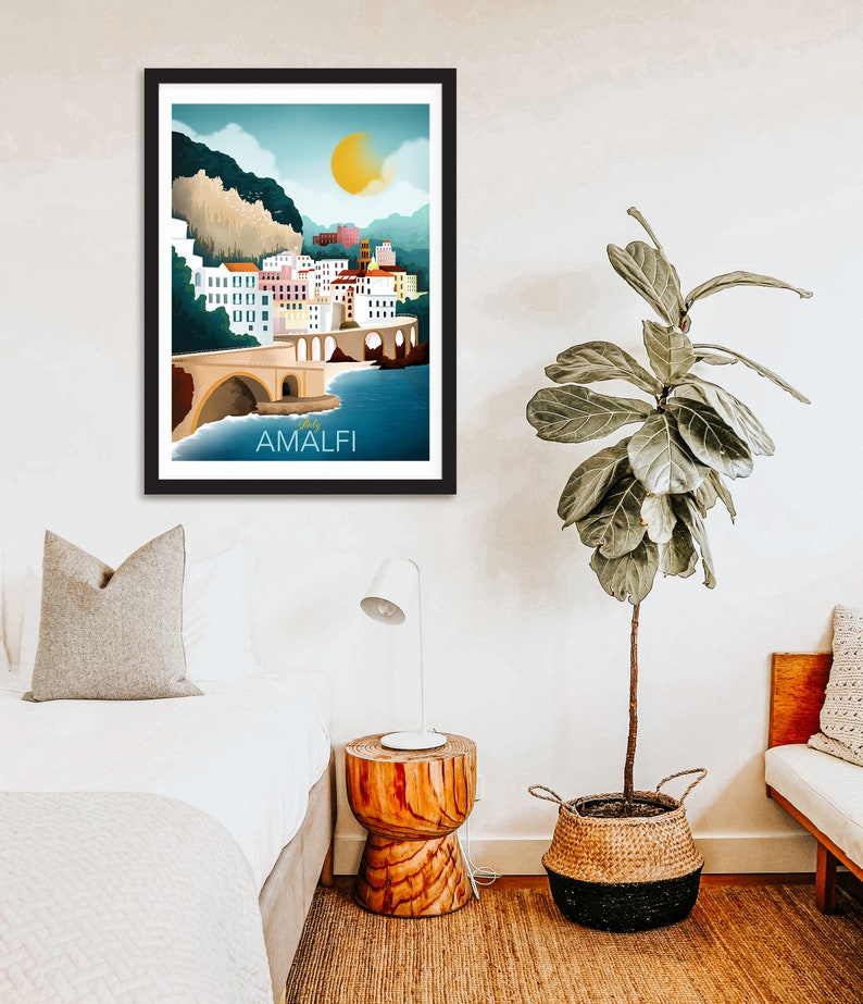 May include: Framed art print featuring a colorful illustration of Amalfi, Italy. The artwork depicts a coastal town with white buildings, a bridge, and the text "AMALFI" in white. The sky is blue with a yellow sun.