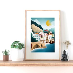 May include: A framed art print depicting a coastal town with white buildings, a bridge, and a bright yellow sun. The artwork features a serene landscape with blue water and a mountain in the background. The frame is made of light-colored wood.