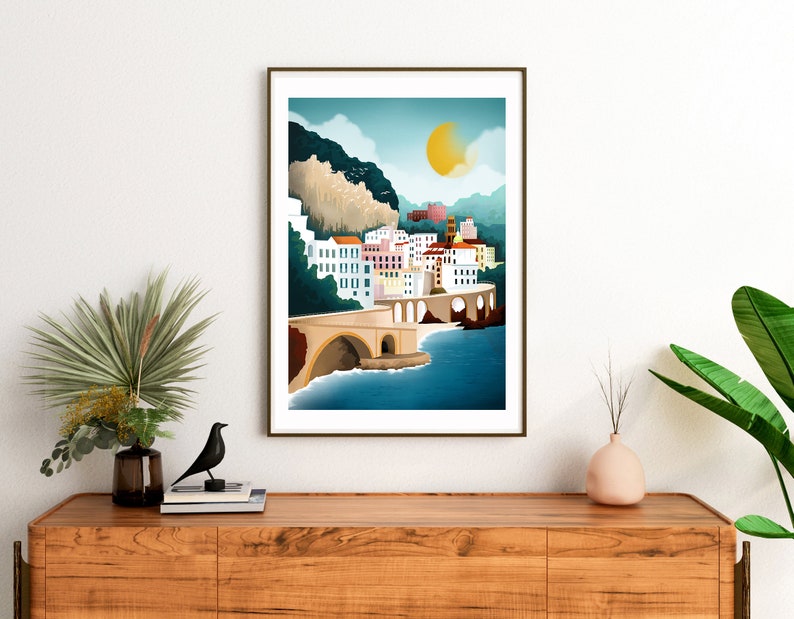 May include: A framed art print depicting a coastal town scene. The artwork features a bridge, buildings with colorful facades, and a large yellow sun in a blue sky. The print is displayed on a wooden cabinet with decorative items.