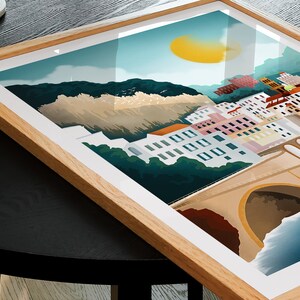 May include: A framed art print featuring a coastal town scene with colorful buildings, a bridge, and a bright yellow sun. The artwork is set against a backdrop of mountains and a blue sky, encased in a light brown wooden frame.