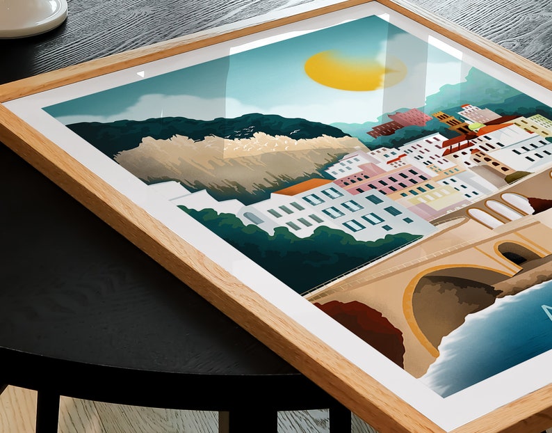 May include: A framed art print featuring a coastal town scene with colorful buildings, a bridge, and a bright yellow sun. The artwork is set against a backdrop of mountains and a blue sky, encased in a light brown wooden frame.