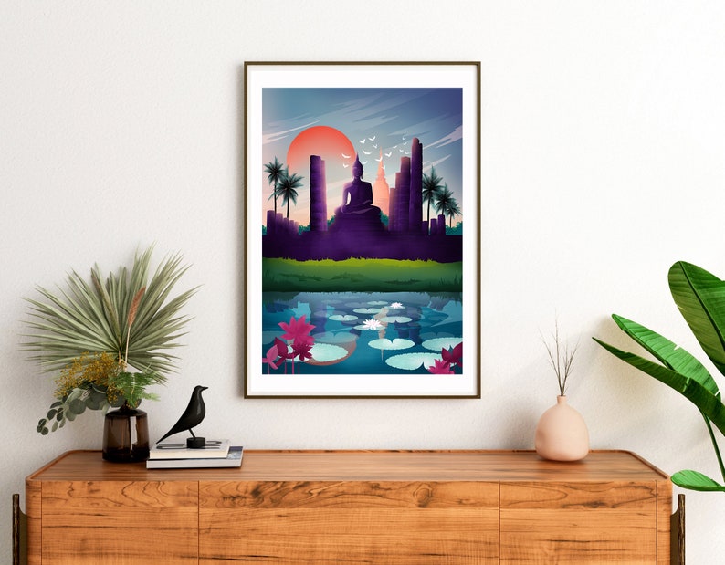 May include: Framed art print showing a tranquil scene with a seated Buddha, temples, and palm trees set against a sunset. A reflective body of water with lily pads and lotus flowers is in the foreground. The artwork is displayed on a wooden cabinet.
