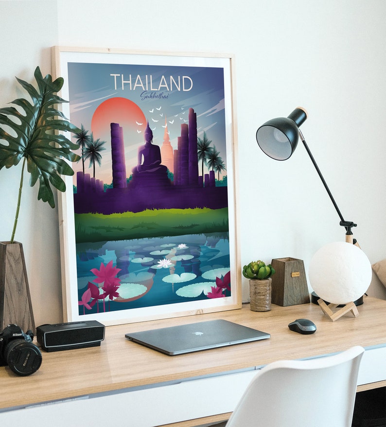 May include: A framed art print of a Thailand Sukhothai landscape. The artwork shows a seated Buddha, temples, and palm trees under a sunset. The print is on a wooden desk with a laptop, lamp, and decorative items.