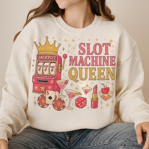 Puede incluir: Sudadera color crema con un gráfico de estilo retro. El diseño presenta una máquina tragamonedas rosa con una corona dorada, el texto "SLOT MACHINE QUEEN" en rosa y dorado, e ilustraciones temáticas de casino.