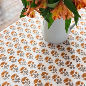 May include: A white tablecloth with a repeating pattern of orange floral designs. A white pitcher holds a bouquet of orange flowers with green leaves. The tablecloth is likely made of cotton or linen.