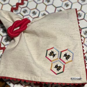 May include: A beige linen napkin with a red knotted tie and embroidered bees in a honeycomb pattern. The napkin has a red scalloped edge and a small white label with the text "MAM EG" on it.