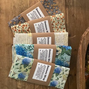 May include: A collection of colorful beeswax wraps with floral patterns, including blue hydrangeas and yellow flowers. Each wrap is packaged with a brown paper label that reads "Beeswax Wrap". The wraps are arranged on a wooden surface.