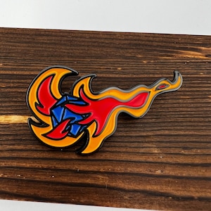 May include: A blue and red enamel pin with a stylized flame design. The flame is yellow and orange and surrounds a blue diamond shape.