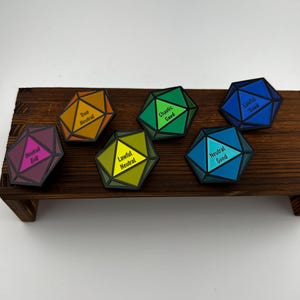 May include: Seven colorful, geometric dice with labels like "True Neutral" and "Chaotic Good" are arranged on a small wooden shelf. Each die has a different color scheme, including orange, green, and blue, with white text.