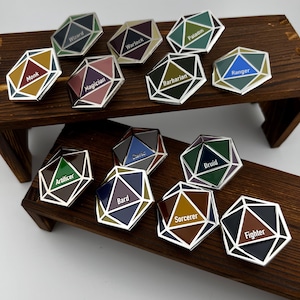 May include: A set of 12 Dungeons and Dragons character class enamel pins. Each pin is shaped like a 20-sided die and features a different class name and color scheme. The classes include: Barbarian, Bard, Cleric, Druid, Fighter, Monk, Paladin, Ranger, Sorcerer, Warlock, Wizard, and Artificer.