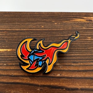 May include: A black enamel pin with a red and yellow flame design. The flame surrounds a blue diamond shape.