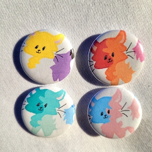 May include: Four round button pins with cartoon cat illustrations. Each pin features a different color combination: yellow and purple, orange and pink, blue and green, and pink and blue.