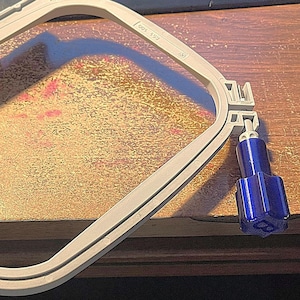 May include: A white plastic embroidery hoop with a blue plastic knob. The hoop is designed for use with an embroidery machine.