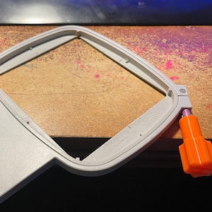 May include: A white plastic embroidery hoop with a 100x100mm marking. The hoop is attached to a white plastic handle with an orange knob.