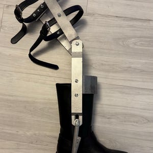 May include: A silver metal leg brace with straps attached to a black leather boot. The brace has a hinged joint and a lever.