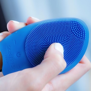 May include: A blue facial cleansing device with a textured silicone surface and the text "SKIN FRESH PARIS". The device has a power button and a plus and minus button. The device is held in a hand.