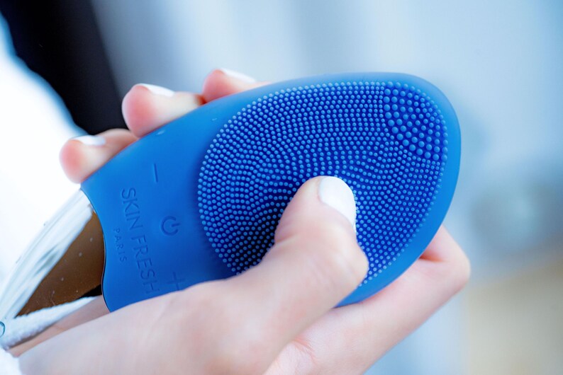 May include: A blue facial cleansing device with a textured silicone surface and the text "SKIN FRESH PARIS". The device has a power button and a plus and minus button. The device is held in a hand.