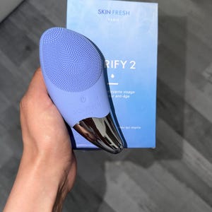 May include: A light purple facial cleansing device with a silver base and the text "SKIN FRESH PARIS". The device is held next to a blue box with the text "RIFY 2".