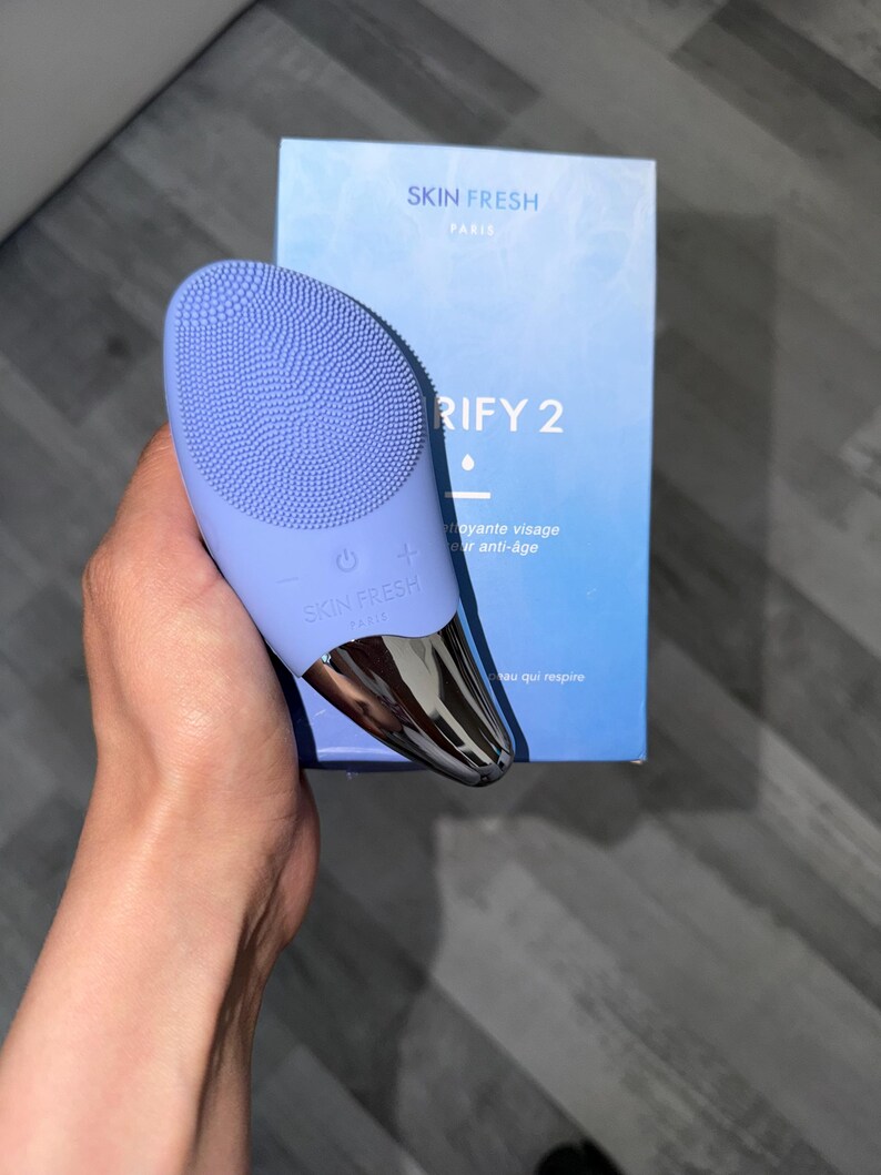 May include: A light purple facial cleansing device with a silver base and the text "SKIN FRESH PARIS". The device is held next to a blue box with the text "RIFY 2".