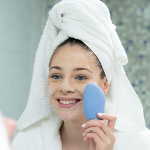 May include: A woman in a white robe and towel uses a blue facial cleansing device. The woman is smiling, and her face is covered in a white cleanser. The background is a bathroom with a patterned tile wall.