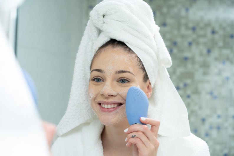 May include: A woman in a white robe and towel uses a blue facial cleansing device. The woman is smiling, and her face is covered in a white cleanser. The background is a bathroom with a patterned tile wall.
