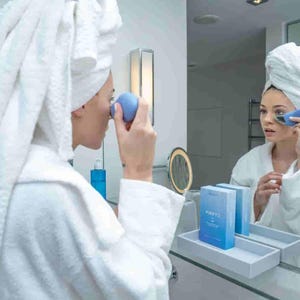 May include: A person in a white robe and towel uses a blue facial device in front of a mirror. The device is held near the eye area. A tray with two blue boxes is visible on the counter.