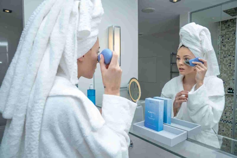 May include: A person in a white robe and towel uses a blue facial device in front of a mirror. The device is held near the eye area. A tray with two blue boxes is visible on the counter.