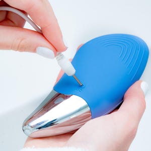 May include: A blue and silver electronic device with a charging port is being plugged in. The device has a smooth, curved design with a silver base and a blue top. The charging cable is white.