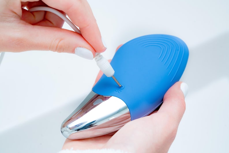 May include: A blue and silver electronic device with a charging port is being plugged in. The device has a smooth, curved design with a silver base and a blue top. The charging cable is white.