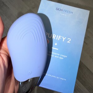 May include: A light blue facial cleansing device with a silver base, next to a blue box. The box reads "SKIN FRESH PARIS PURIFY 2" and includes French text. The device has a DC symbol.
