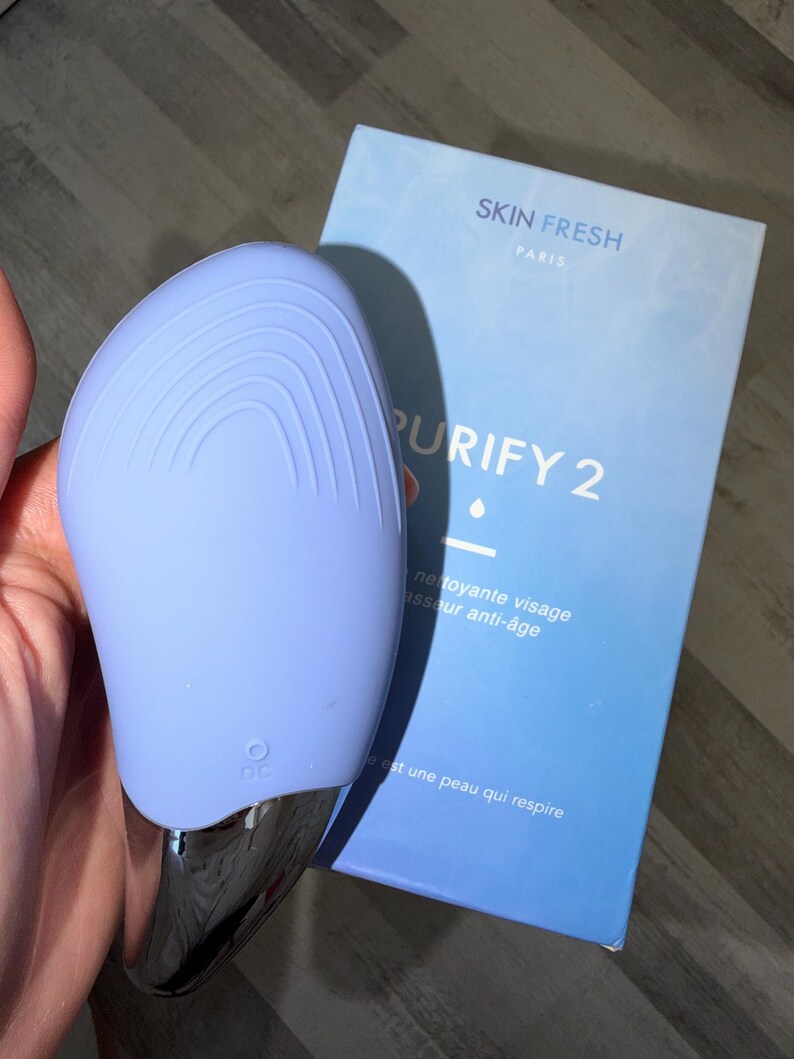May include: A light blue facial cleansing device with a silver base, next to a blue box. The box reads "SKIN FRESH PARIS PURIFY 2" and includes French text. The device has a DC symbol.