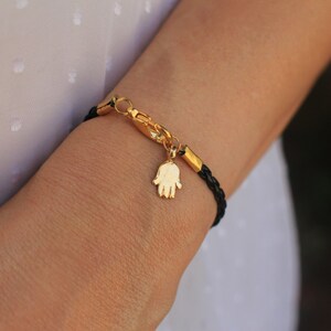 May include: A black leather bracelet with a gold hamsa charm. The bracelet is adjustable and has a lobster clasp closure.