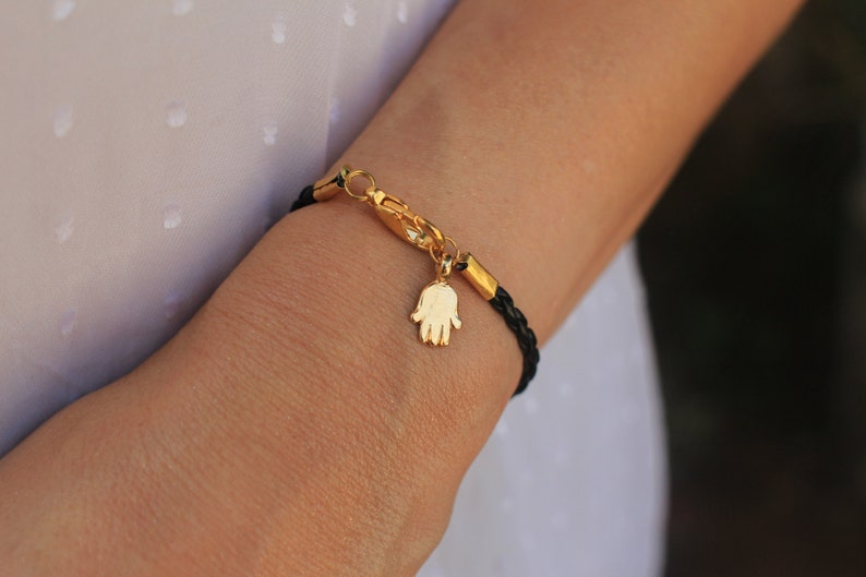 May include: A black leather bracelet with a gold hamsa charm. The bracelet is adjustable and has a lobster clasp closure.